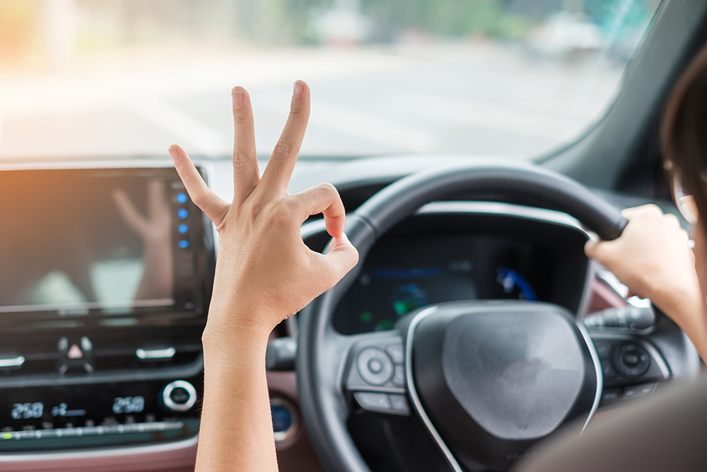 woman showing OK sign during driving a car on the road, hand controlling steering wheel in electric modern automobile. Journey, trip and safety Transportation concepts